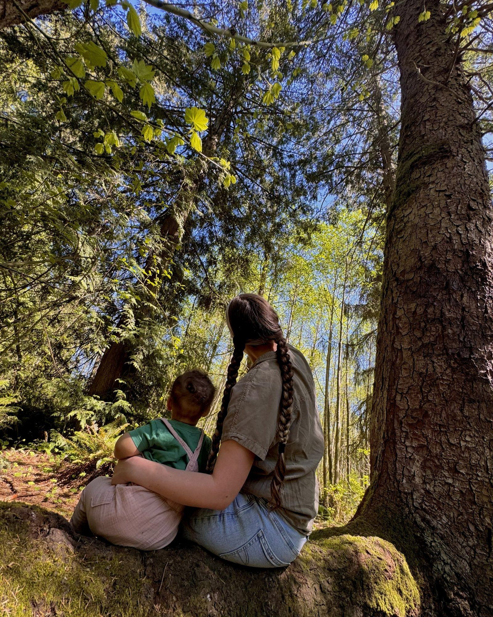 Parent and toddler sitting on a mossy log beneath tall trees, looking up at the forest canopy