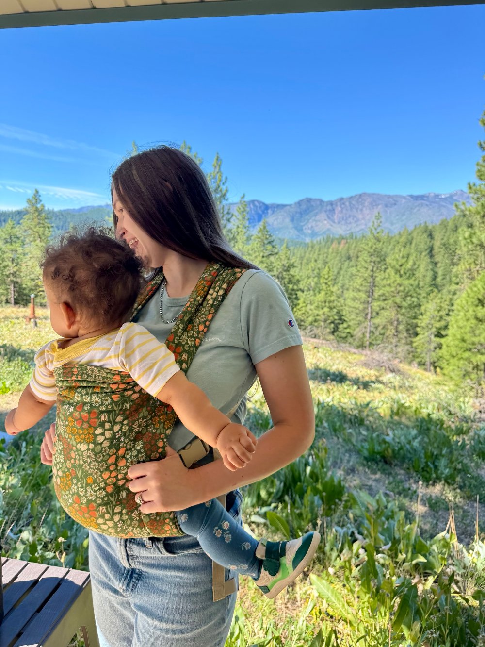 Mattison laughing with her daughter in a baby carrier, with pine trees and mountains behind them