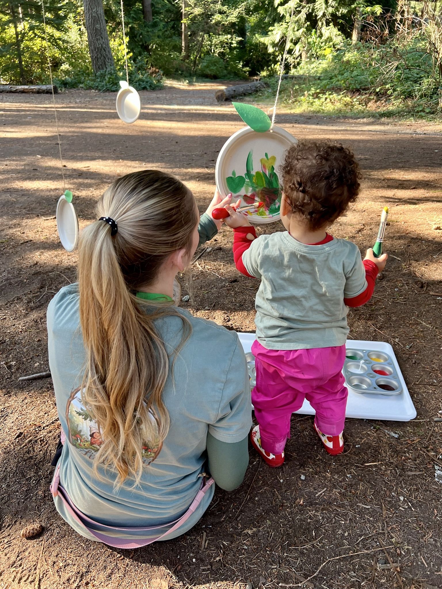 A teacher sits beside a toddler as she paints a paper plate apple hanging from a string in the forest