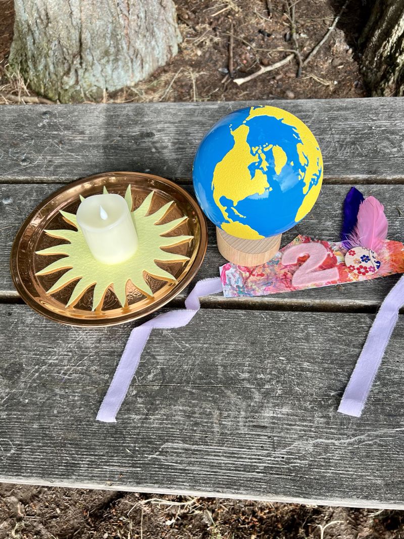 A birthday setup on a picnic table: a candle on a paper sun, a small globe, and a painted birthday crown with a pink number 2, feathers, and lavender ribbon ties
