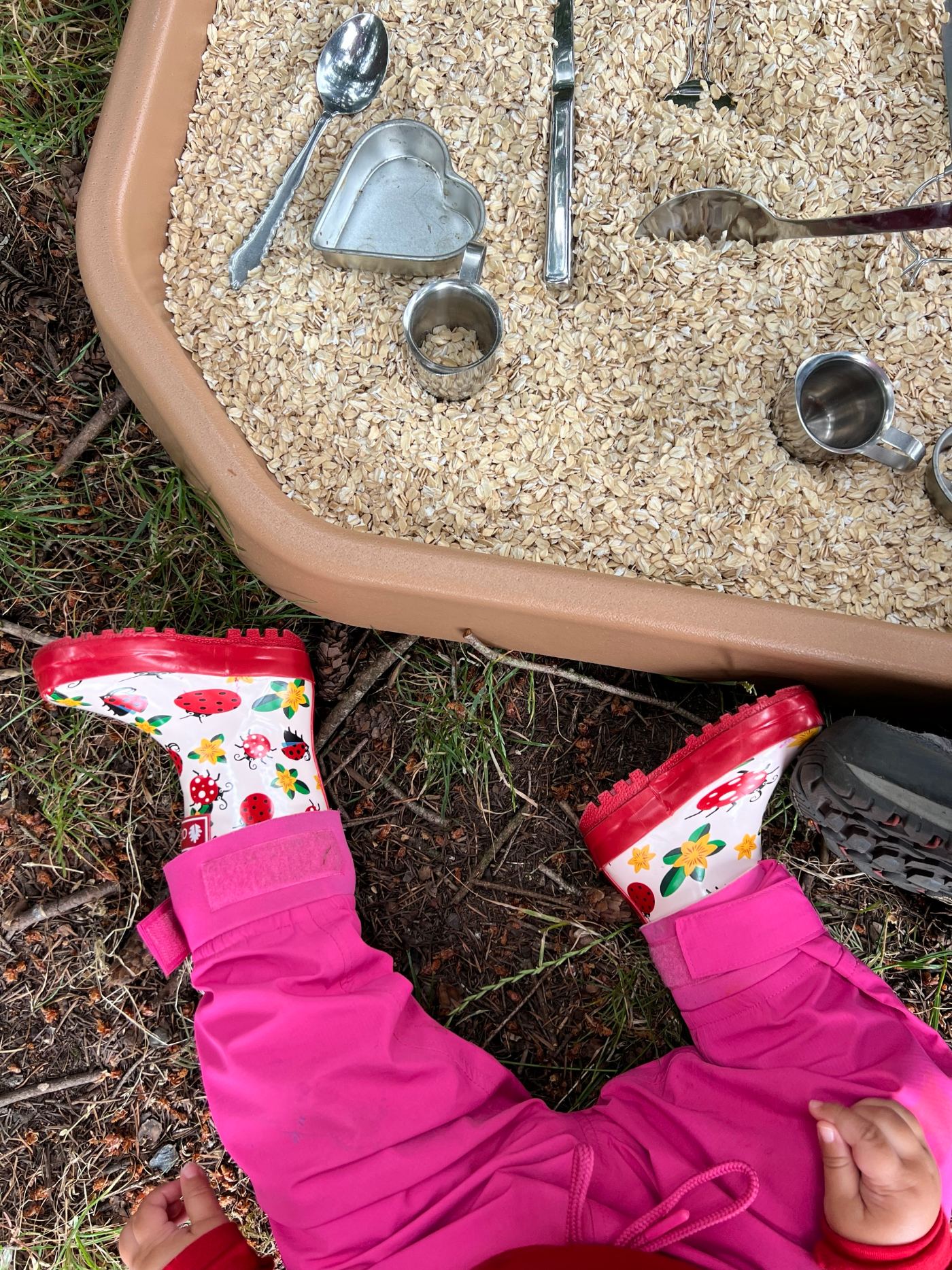 Toddler in ladybug rain boots sitting next to a sensory bin of oats with spoons and cups
