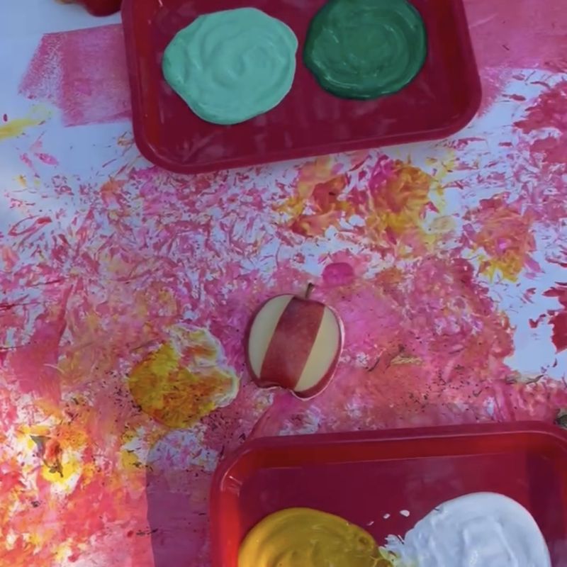 Green and white paint stamped with apple halves over a pink and yellow base on the class canvas