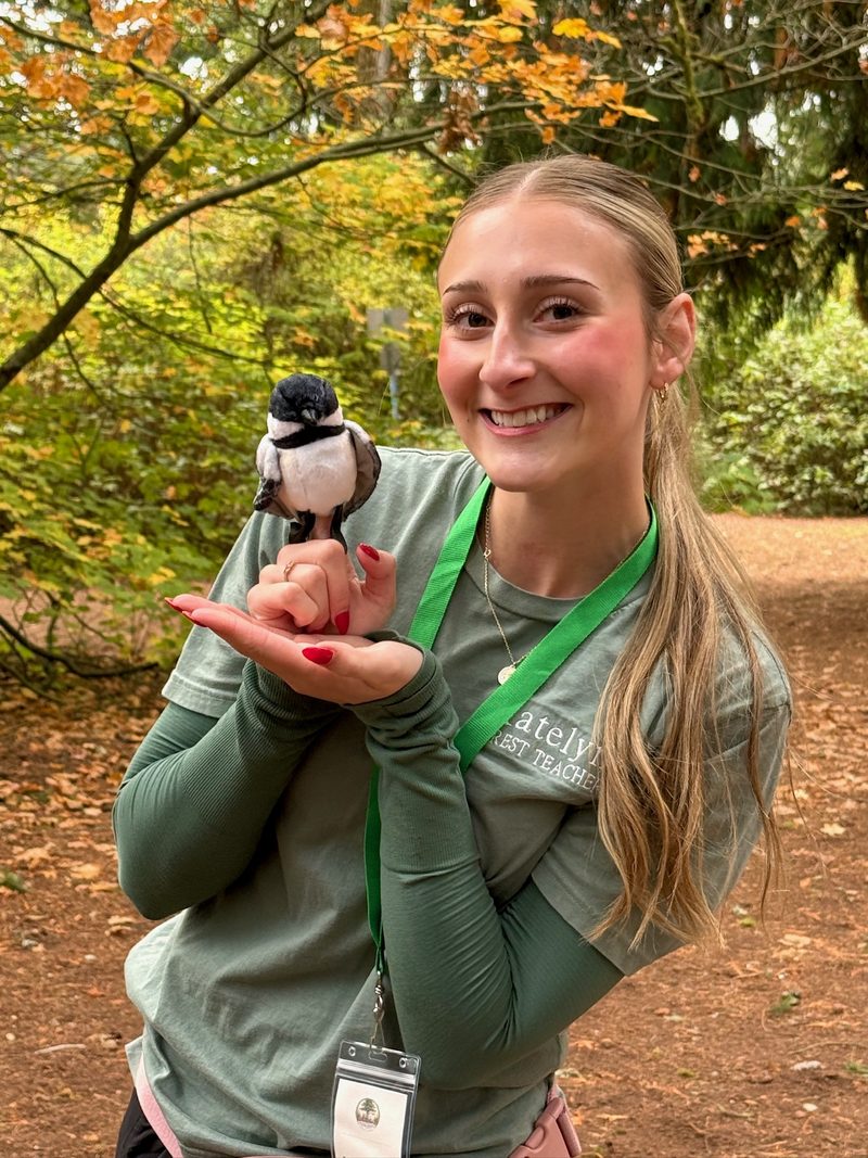 Katelyn smiling on the forest trail in her Forest Teacher shirt, holding Pip the chickadee puppet on her hand, with fall leaves behind her
