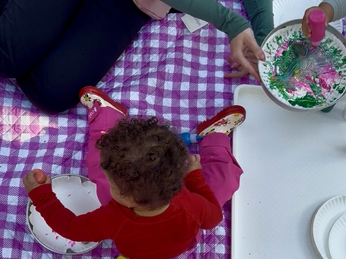 A toddler painting a paper plate while a teacher helps spin another plate of pink and green paint