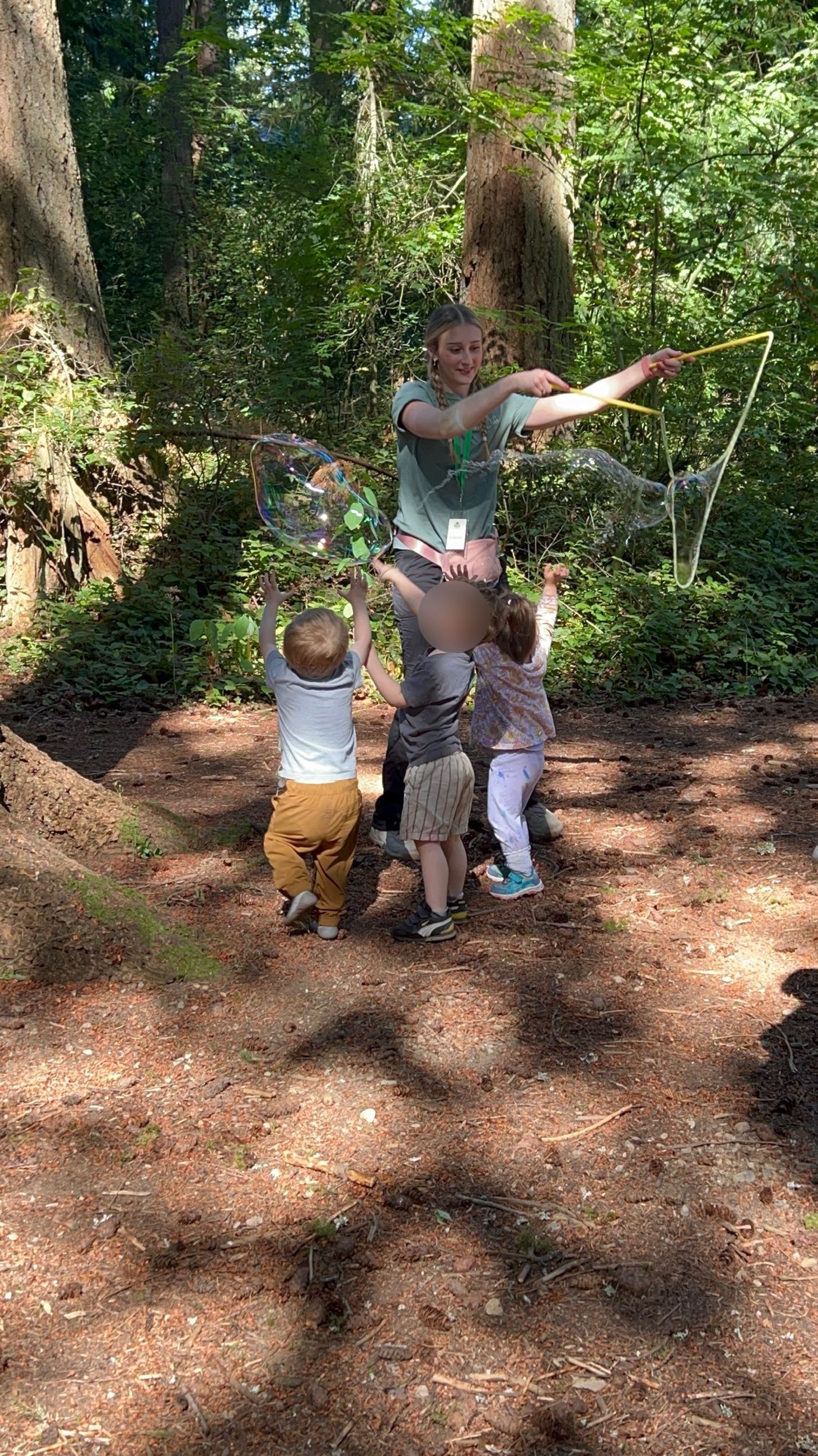 Toddlers reach up to catch giant bubbles a teacher is making in the forest