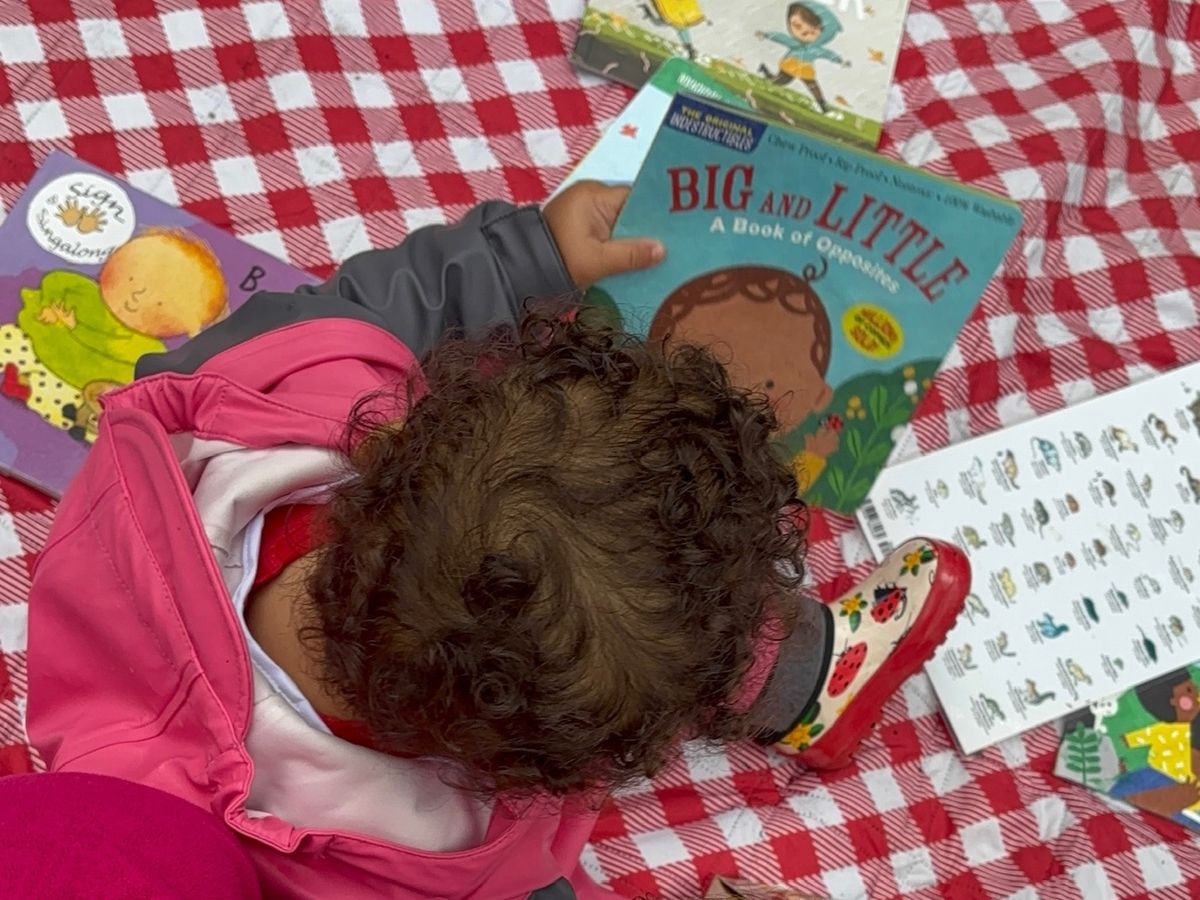 A toddler in a rain suit reading board books on a red checkered blanket