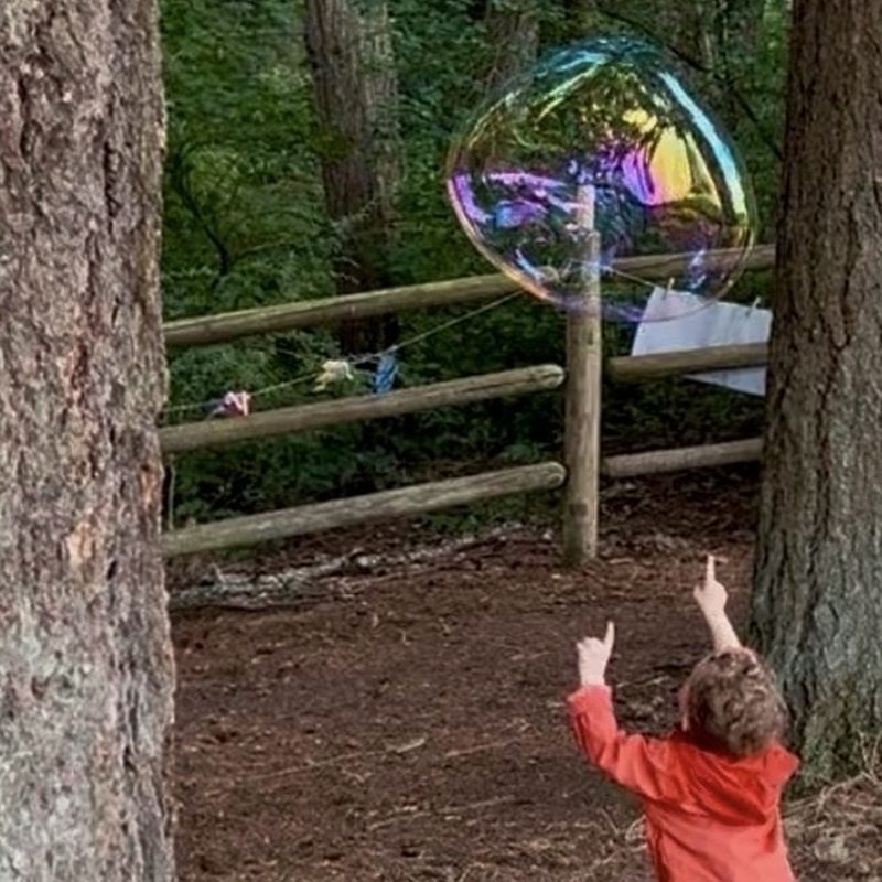 A toddler in rain boots running and reaching for a giant bubble in the forest