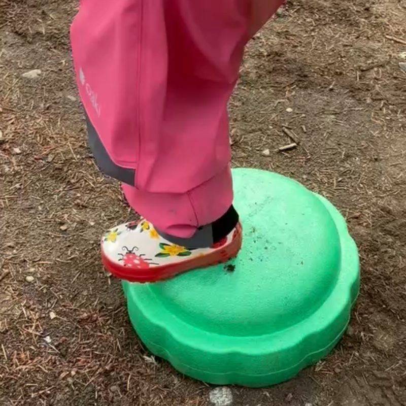 A toddler in a pink rain suit and ladybug boots stepping onto a green pebblestone