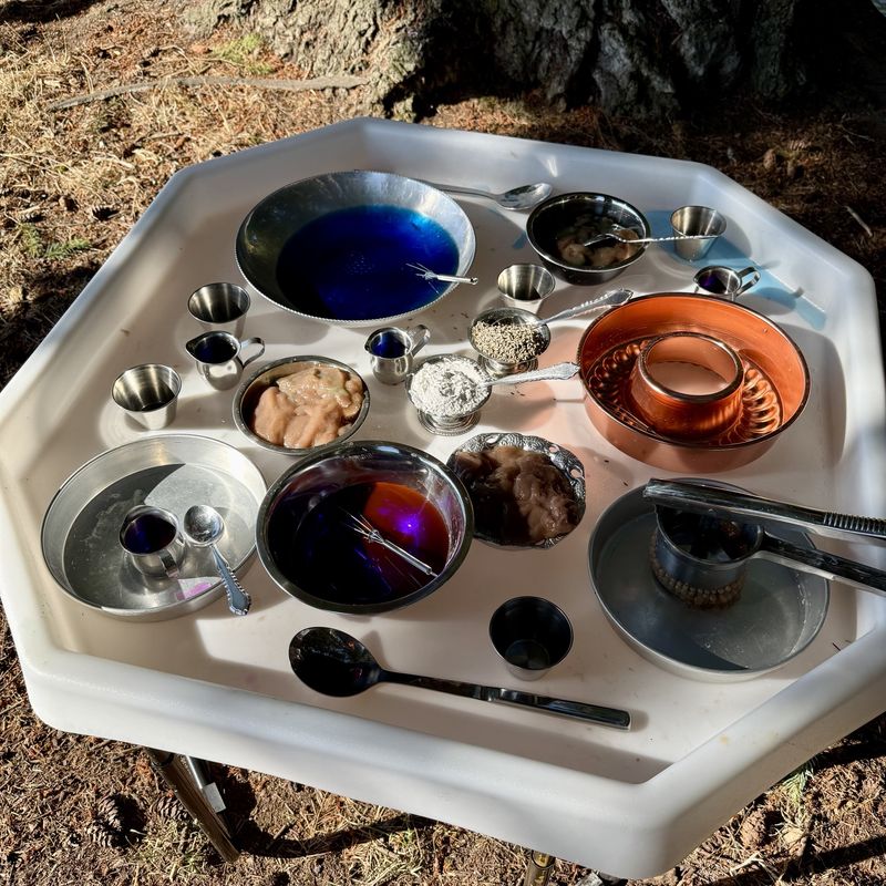 A mud kitchen table under a big tree with blue water, oat mush, flour, a copper mold, and metal bowls and spoons