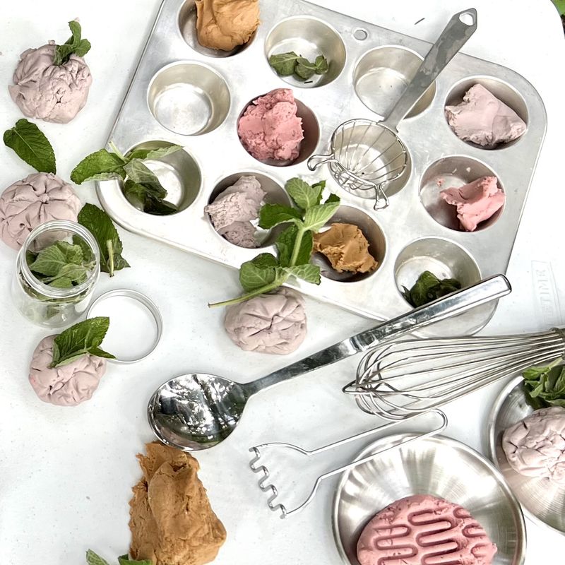 Pink and brown playdough with fresh mint leaves, a muffin tin, whisk, and spoons