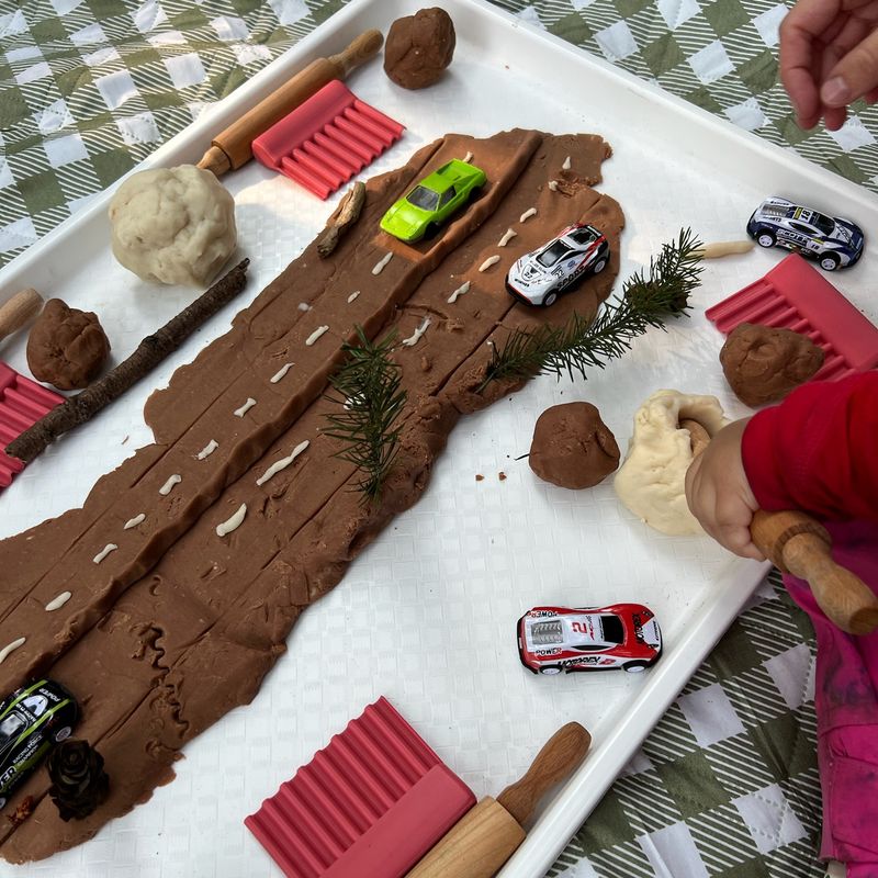 A toddler rolling brown playdough into a road for toy cars, with sticks and pine sprigs