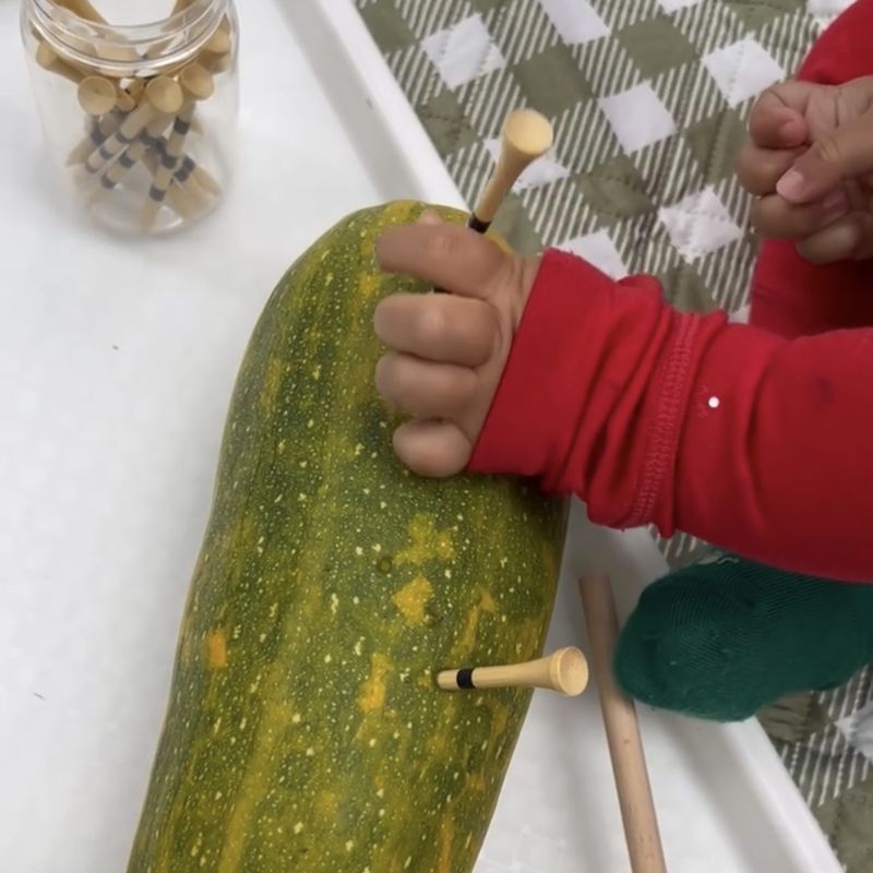 A toddler pushing golf tees into a big squash, with a jar of tees nearby