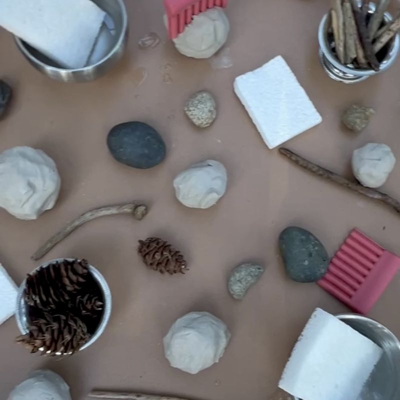 A clay tray with balls of clay, stones, pinecones, sticks, sponges, texture combs, and washcloths