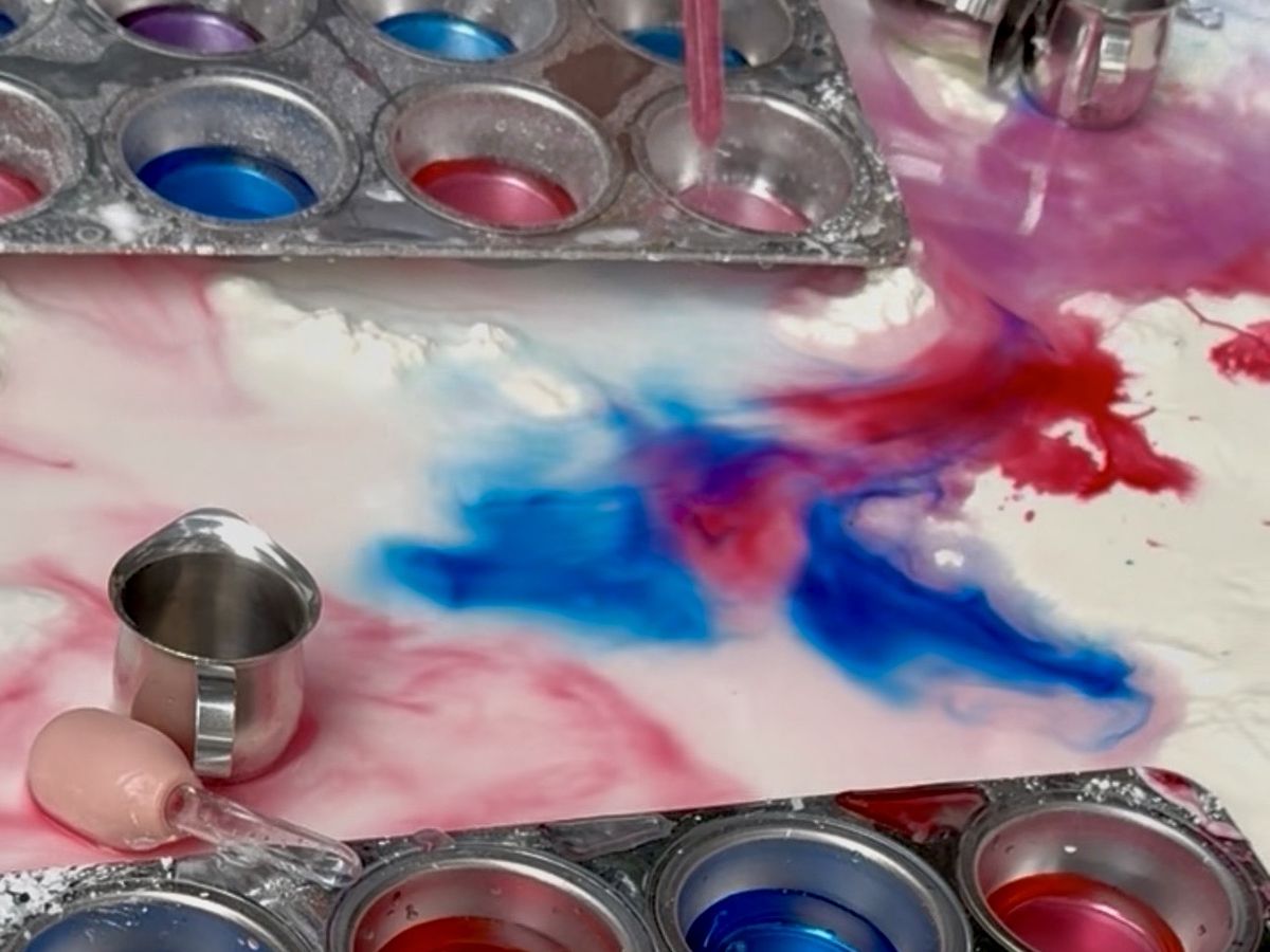 Droppers swirling red and blue colored water into a white tray, with muffin tins of color