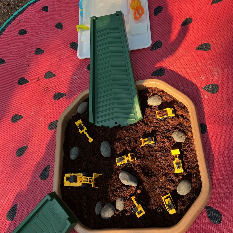 A dig bin full of soil, smooth stones, and toy construction trucks with a chute leading into it