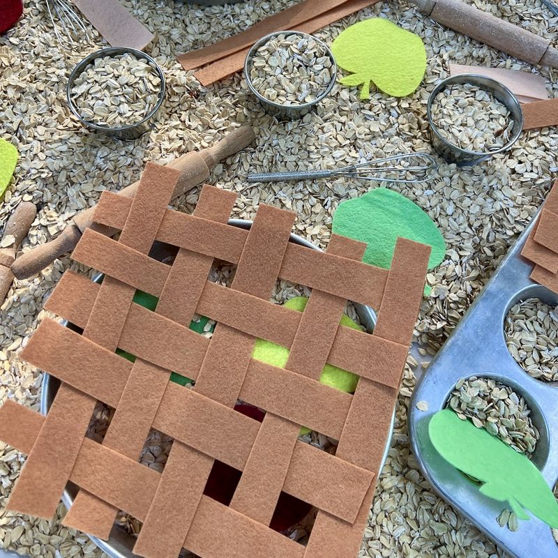 An apple pie sensory bin with oats, felt apples, a felt lattice crust, tiny rolling pins, and tins